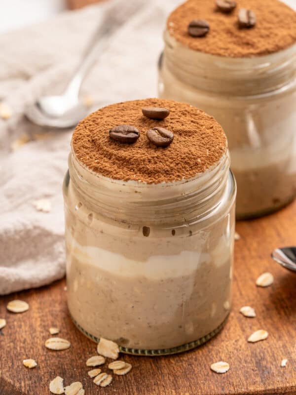 Two glass jars filled with creamy tiramisu overnight oats, topped with a layer of cocoa powder and coffee beans, are placed on a wooden surface with scattered oat flakes and a spoon nearby.
