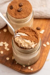 A jar of creamy dessert topped with cocoa powder, with a spoon scooping out a portion. Another jar sits behind it, both on a wooden board sprinkled with oats and coffee beans.