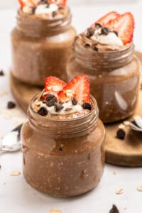 Three jars of chocolate overnight oats are topped with sliced strawberries, chocolate chips, oats, and a dollop of yogurt or cream. The jars sit on a wooden tray with spoons nearby.