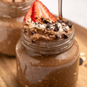 A small glass jar filled with chocolate overnight oats, topped with sliced strawberries, whipped cream, chocolate shavings, and oats. A spoon is inserted into the jar. Another similar jar is in the background.