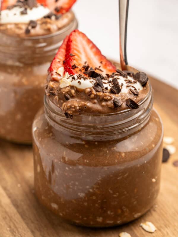 A small glass jar filled with chocolate overnight oats, topped with sliced strawberries, whipped cream, chocolate shavings, and oats. A spoon is inserted into the jar. Another similar jar is in the background.