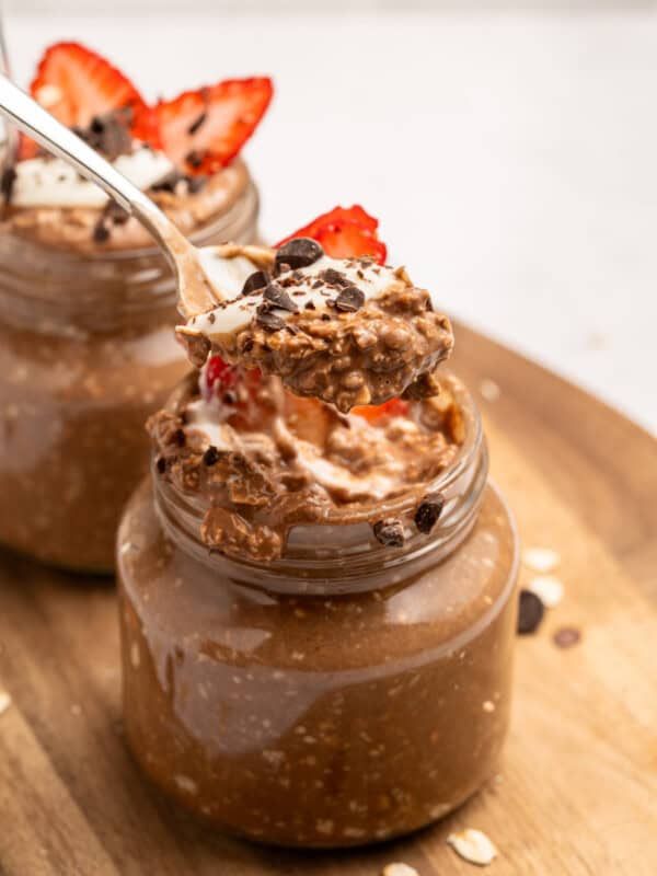 A spoonful of chocolate overnight oats with strawberry slices is held above a glass jar filled with more oats, topped with chocolate chips and a strawberry, on a wooden surface.