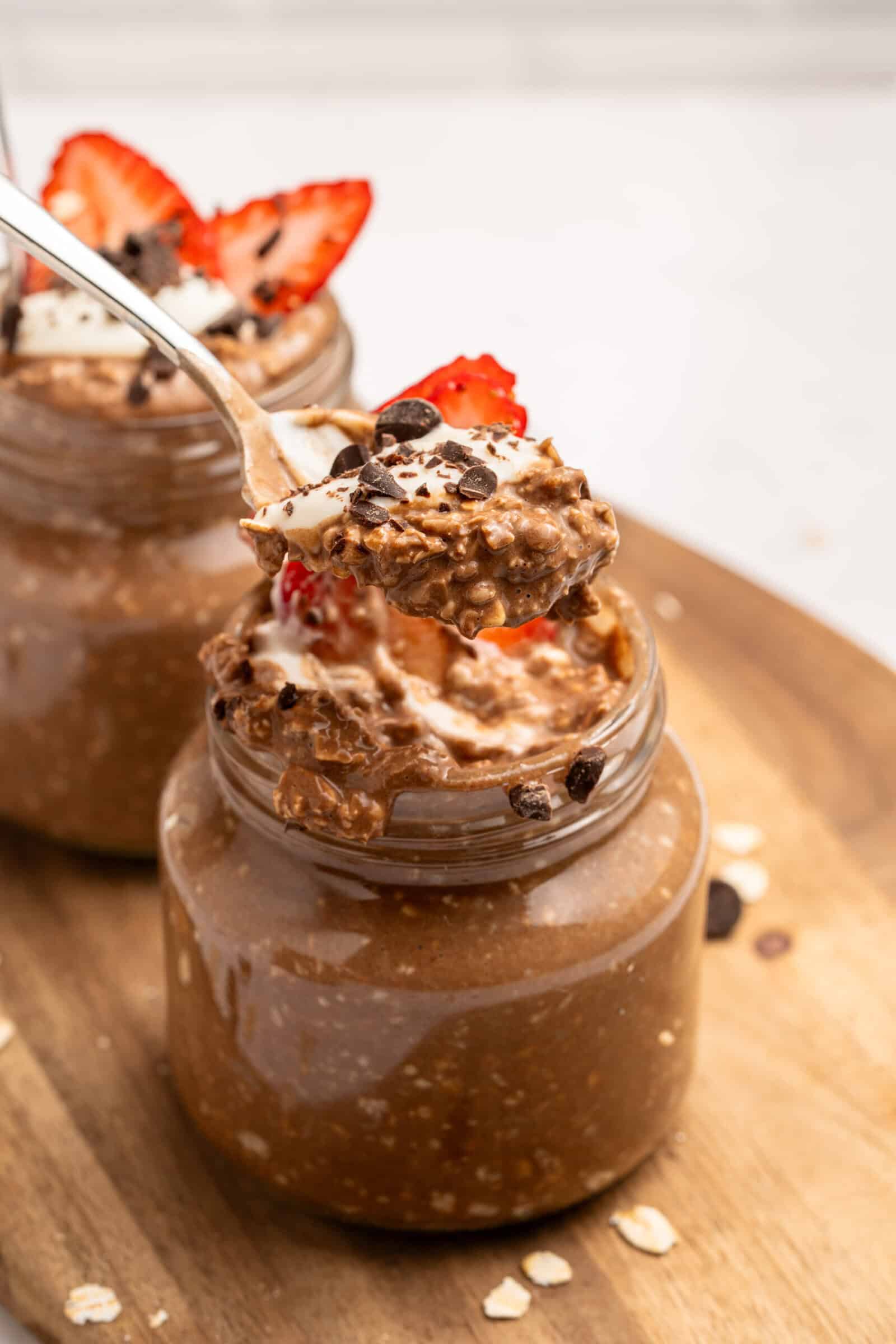 A spoonful of chocolate overnight oats with strawberry slices is held above a glass jar filled with more oats, topped with chocolate chips and a strawberry, on a wooden surface.