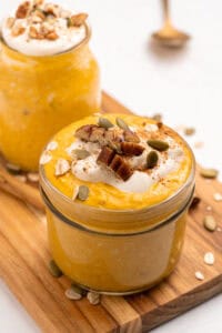 A glass jar filled with creamy orange pumpkin overnight oats, topped with yogurt, pecans, pumpkin seeds, oats, and a sprinkle of cinnamon, sits on a wooden board next to another similar jar.