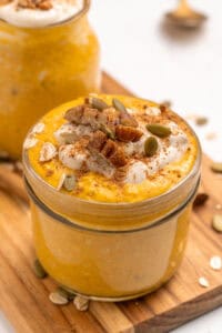 A glass jar filled with creamy pumpkin overnight oats, topped with whipped cream, pecans, pumpkin seeds, oats, and a sprinkle of cinnamon, sitting on a wooden board. Another similar jar is in the background.