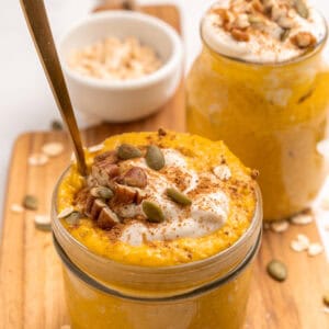 Two glass jars filled with creamy pumpkin overnight oats, topped with yogurt, pecans, pumpkin seeds, and a sprinkle of cinnamon. A spoon is in one jar. Rolled oats are scattered on the wooden board underneath.
