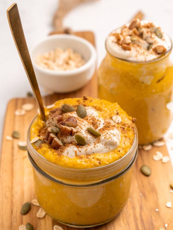 Two glass jars filled with creamy pumpkin overnight oats, topped with yogurt, pecans, pumpkin seeds, and a sprinkle of cinnamon. A spoon is in one jar. Rolled oats are scattered on the wooden board underneath.