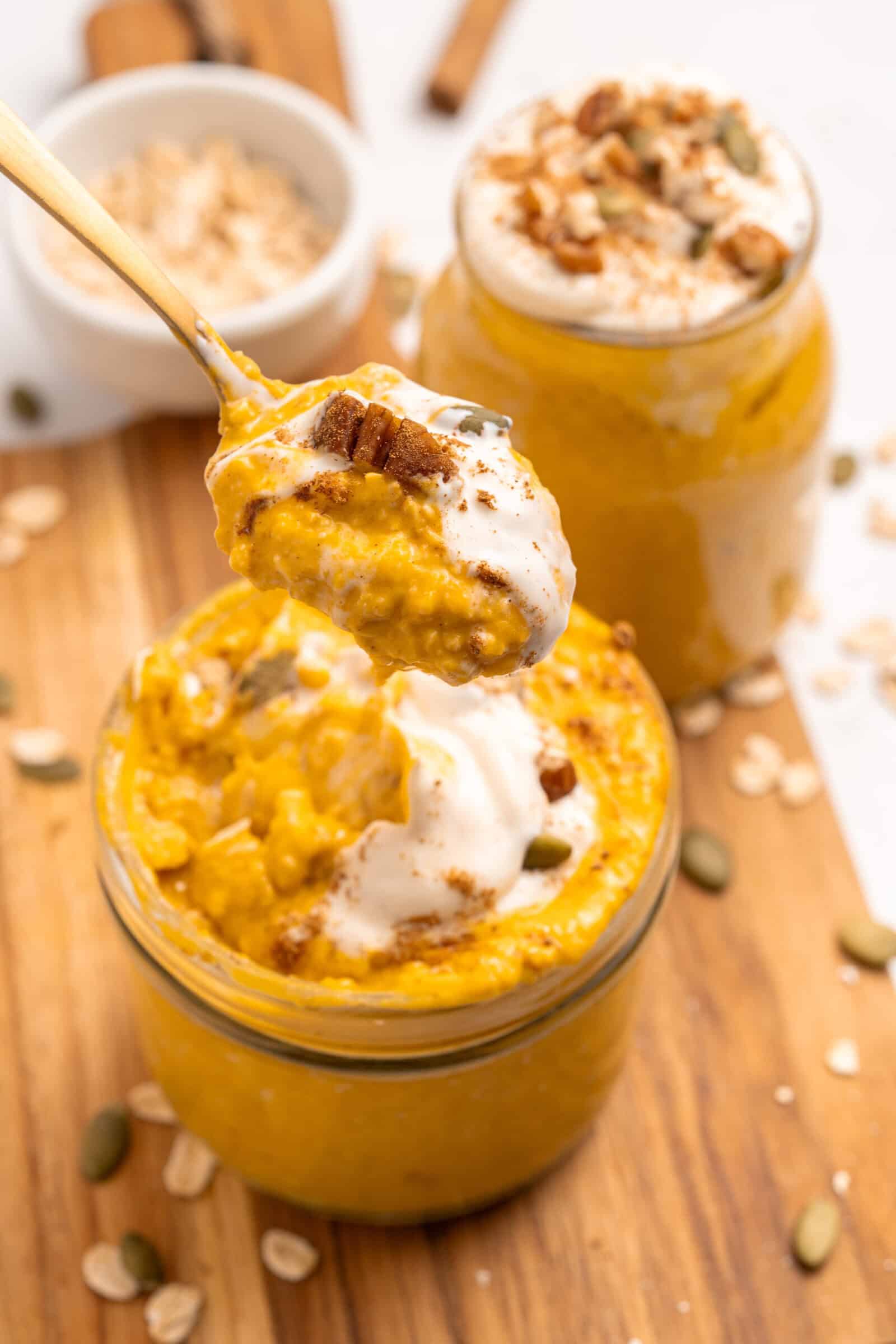 A close-up of pumpkin overnight oats in a jar, topped with cream and nuts, with a spoon lifting a portion. Another jar of oats and scattered oats and seeds are visible on a wooden surface.