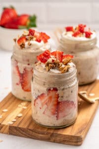 Three glass jars filled with overnight oats, layered with fresh strawberry slices and topped with granola and strawberry pieces, are arranged on a wooden board. A bowl of whole strawberries is in the background.