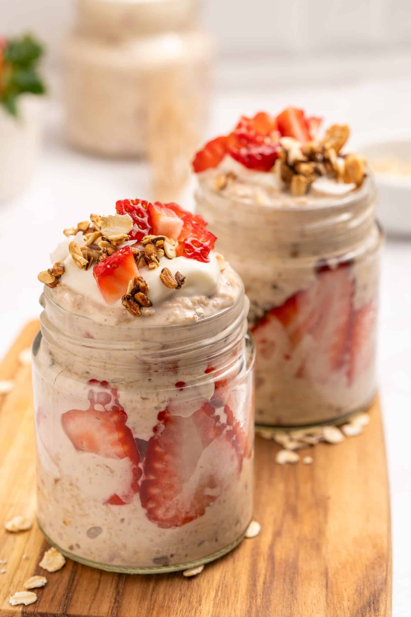 Two glass jars filled with overnight oats, layered with sliced strawberries and topped with creamy yogurt, fresh strawberry pieces, and granola, placed on a wooden board.