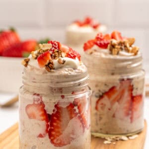 Two glass jars filled with overnight oats, layered with sliced strawberries, topped with granola and whipped cream, sit on a wooden board with fresh strawberries and oats scattered around.