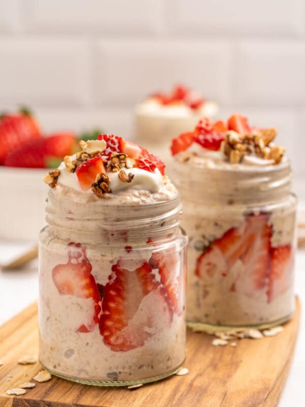 Two glass jars filled with overnight oats, layered with sliced strawberries, topped with granola and whipped cream, sit on a wooden board with fresh strawberries and oats scattered around.