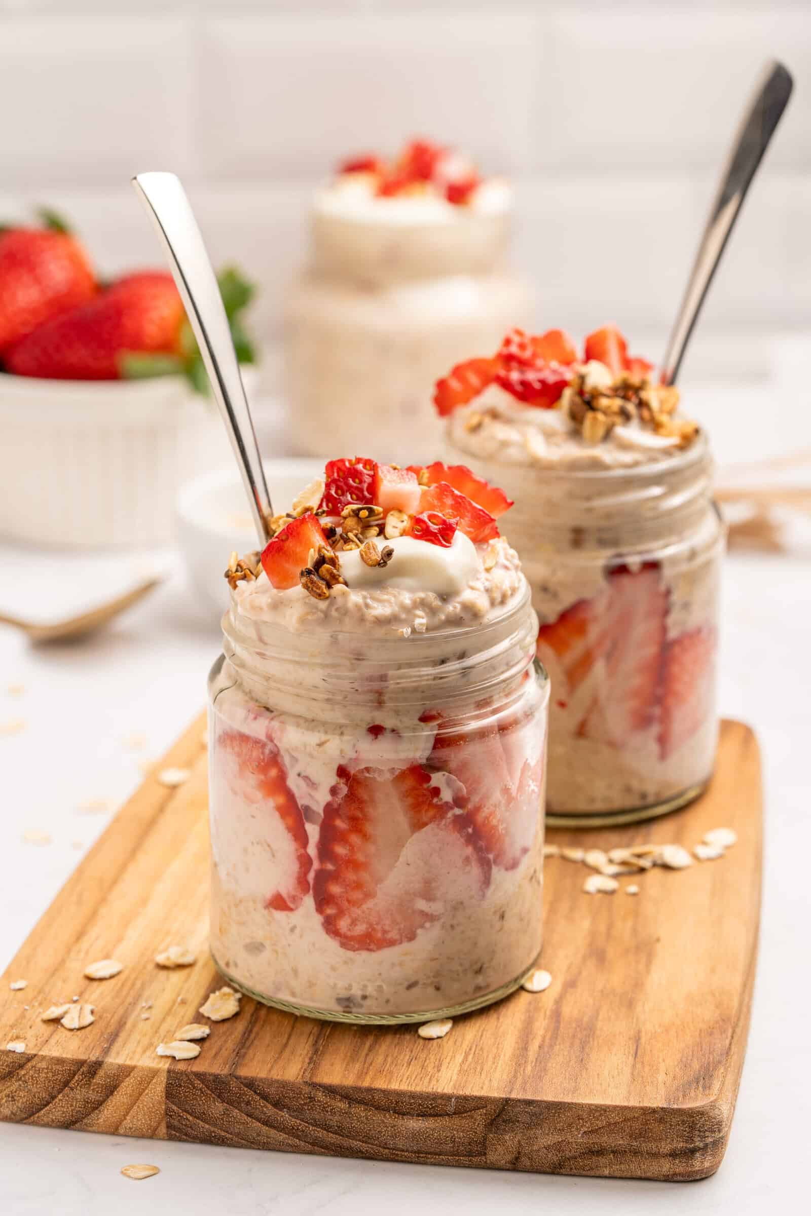 Two glass jars filled and layered with sliced strawberries and topped with granola and yogurt, sit on a wooden board. A bowl of strawberries is in the background, and oats are scattered around.