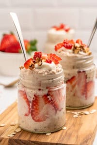 Two glass jars filled with overnight oats, layered with sliced strawberries and topped with granola, yogurt, and more strawberries. Each jar has a spoon inside and sits on a wooden board with oats scattered around.