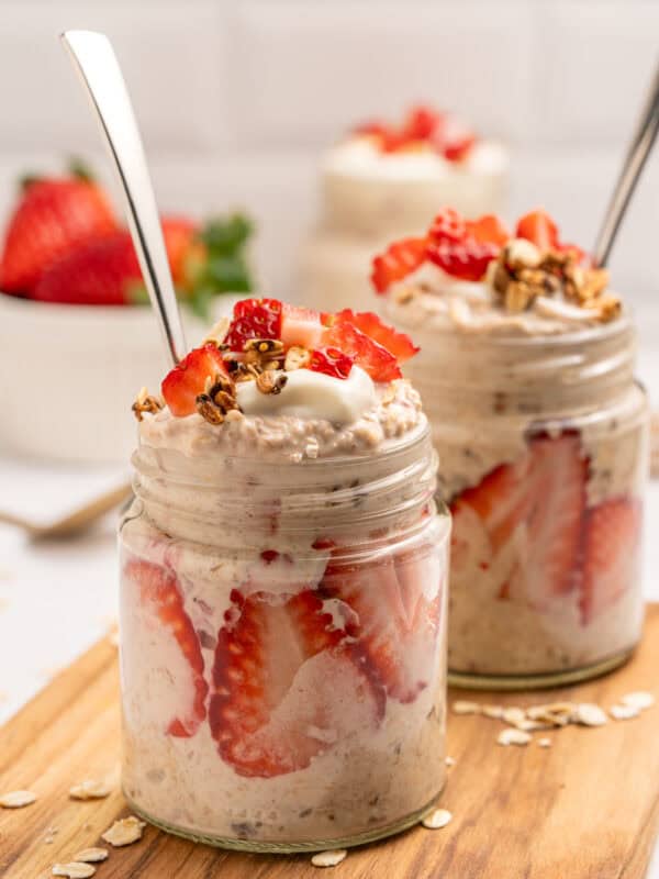 Two glass jars filled with overnight oats, layered with sliced strawberries and topped with granola, yogurt, and more strawberries. Each jar has a spoon inside and sits on a wooden board with oats scattered around.