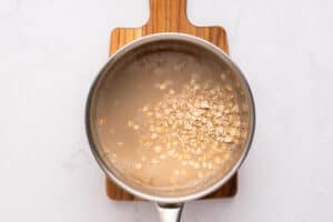 A metal saucepan with oats soaking in water sits on a small wooden cutting board, viewed from above on a white surface.