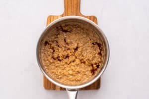 A saucepan filled with cooked oatmeal, topped with a swirl of dark syrup, sits on a small wooden cutting board against a light-colored background.