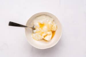 A white bowl with plain yogurt and honey, partially mixed, with a metal spoon resting inside, on a white surface.