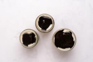Three small glass jars filled with a creamy dessert topped with a layer of glossy dark chocolate sauce, arranged in a triangle on a white marble surface.