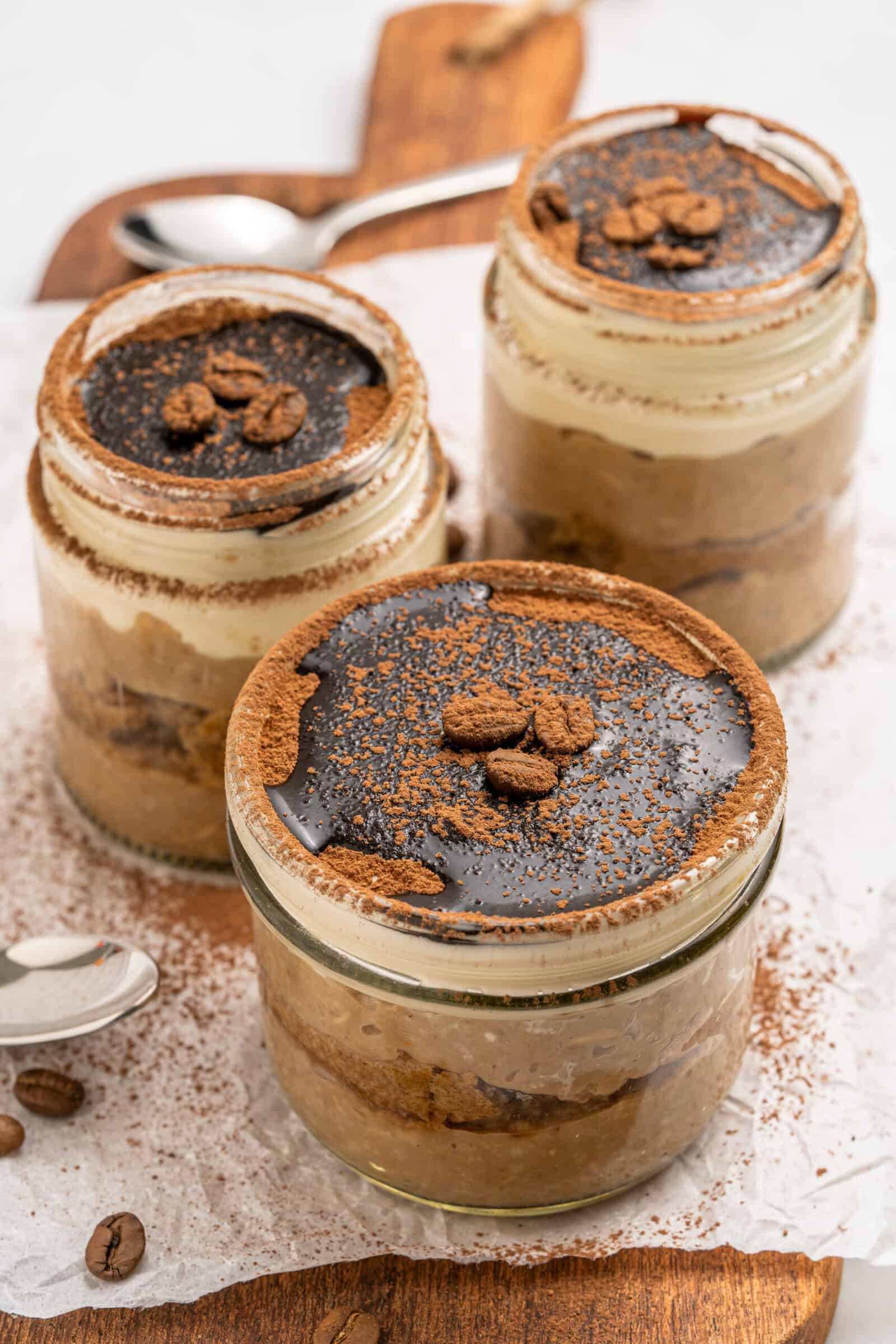 Three glass jars filled with layered tiramisu dessert, topped with cocoa powder, chocolate, and coffee beans, are arranged on a wooden board with a spoon and scattered coffee beans nearby.