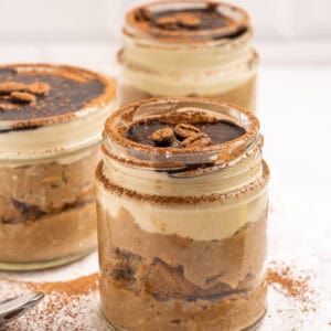 Three jars of tiramisu dessert layered with cream, cake, and cocoa, topped with chocolate sauce and coffee beans, sit on a wooden board with cocoa powder and coffee beans scattered around.