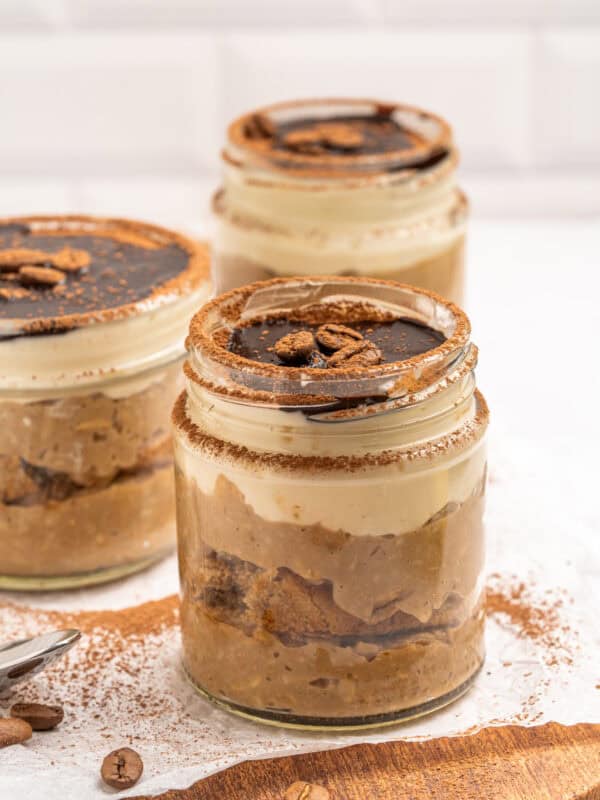 Three jars of tiramisu dessert layered with cream, cake, and cocoa, topped with chocolate sauce and coffee beans, sit on a wooden board with cocoa powder and coffee beans scattered around.