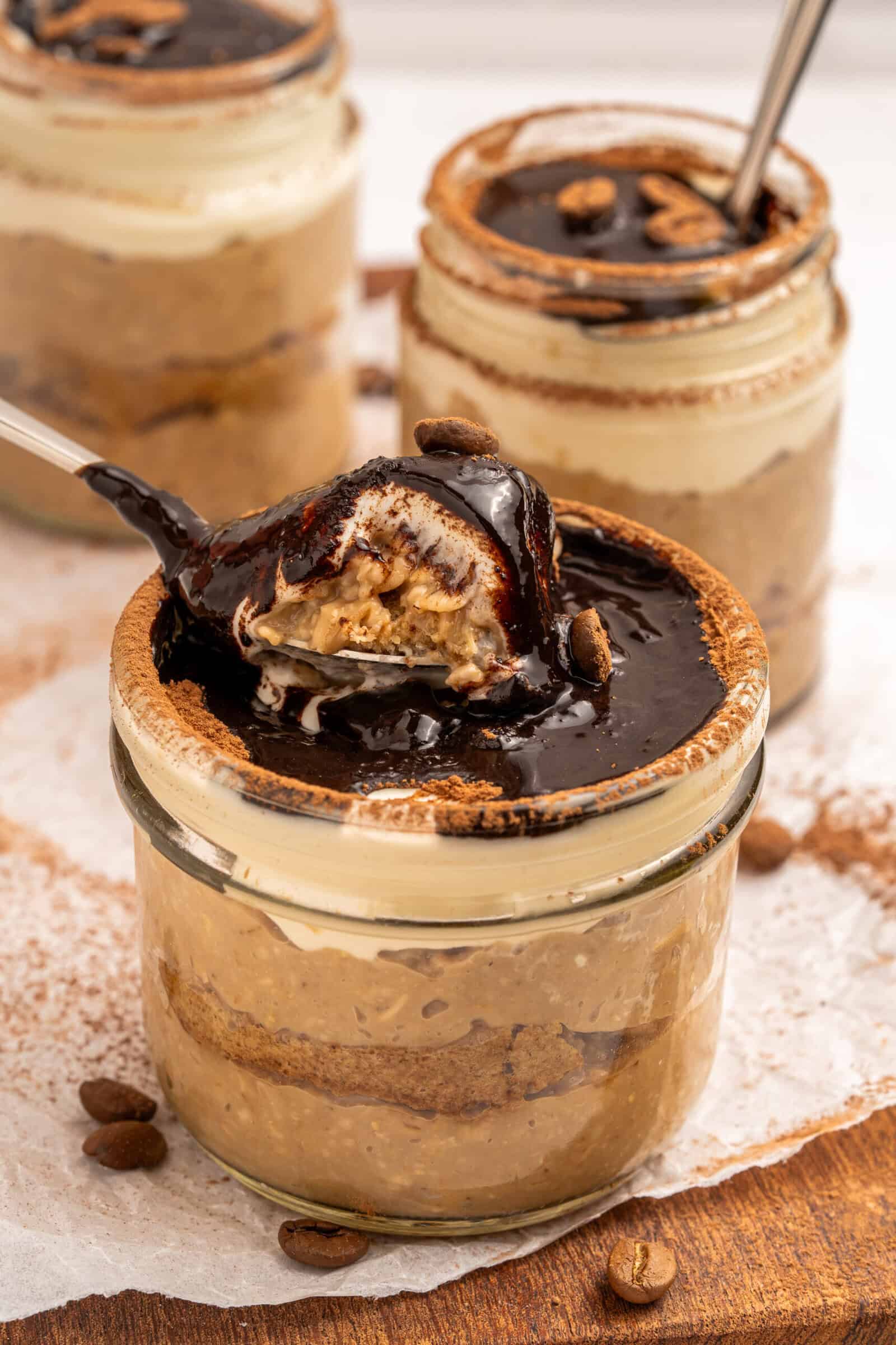 Three glass jars filled with creamy tiramisu, topped with chocolate sauce and coffee beans. One jar has a spoonful lifted, showing layers of dessert inside. The jars rest on parchment paper with scattered coffee beans.