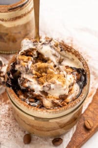 A close-up of a jar filled with creamy tiramisu, topped with swirls of mascarpone, cocoa powder, and coffee-soaked ladyfingers, with a spoon scooping out a portion. Coffee beans are scattered nearby.