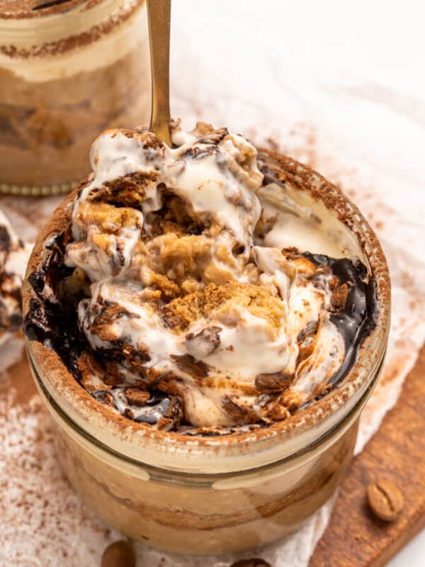 A close-up of a jar filled with creamy tiramisu, topped with swirls of mascarpone, cocoa powder, and coffee-soaked ladyfingers, with a spoon scooping out a portion. Coffee beans are scattered nearby.