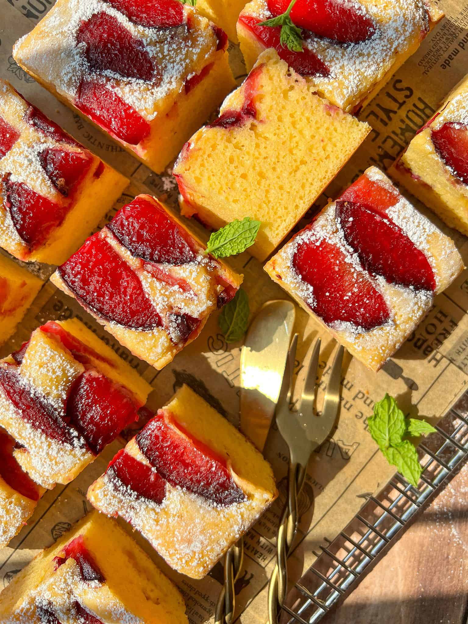 Squares of yellow sponge cake topped with baked red plums and a dusting of powdered sugar are arranged on a cooling rack with vintage paper underneath. Mint leaves and gold dessert forks are also visible.