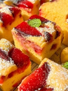Close-up of several pieces of yellow sponge cake topped with red plum slices and powdered sugar, some garnished with a small green mint leaf. The cake pieces are arranged closely together.