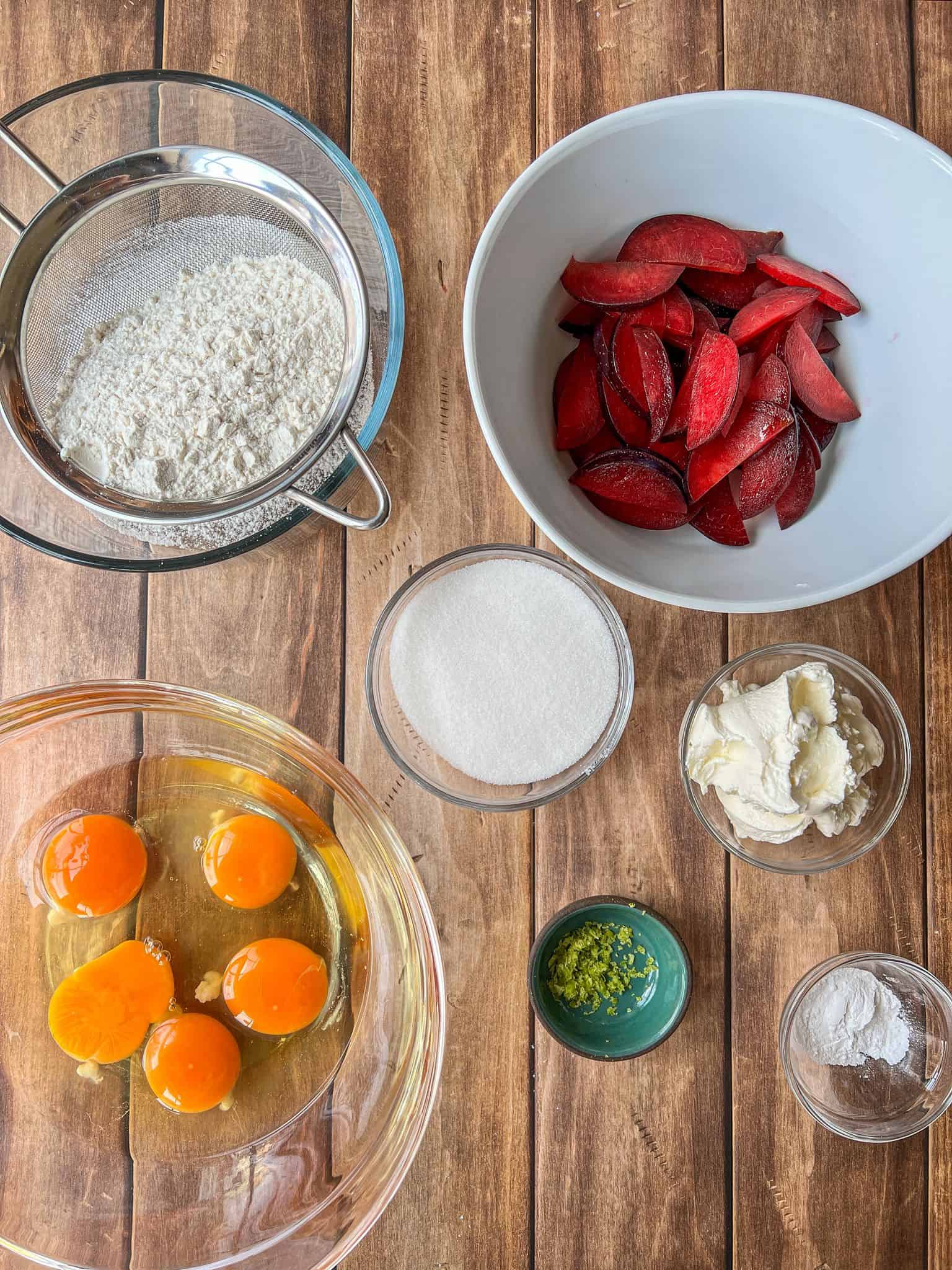 A top-down view of baking ingredients on a wooden surface: sifted flour, sliced plums, eggs in a bowl, a bowl of sugar, a small dish of lime zest, cream cheese, and a small bowl of baking powder.