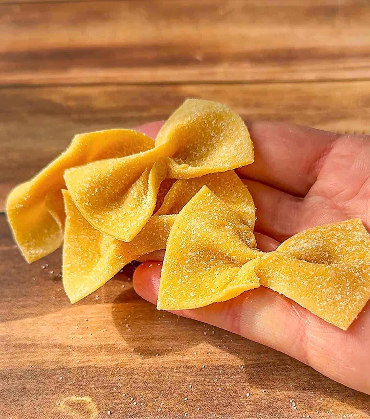 A hand holds three pieces of uncooked farfalle pasta on a wooden surface, with a light dusting of flour visible on the pasta and hand.