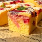 A close-up of a slice of yellow sponge cake topped with red fruit and a dusting of powdered sugar, garnished with a fresh mint leaf, sitting on brown parchment paper with printed text.