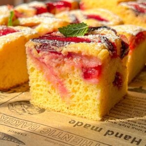 A close-up of a slice of yellow sponge cake topped with red fruit and a dusting of powdered sugar, garnished with a fresh mint leaf, sitting on brown parchment paper with printed text.