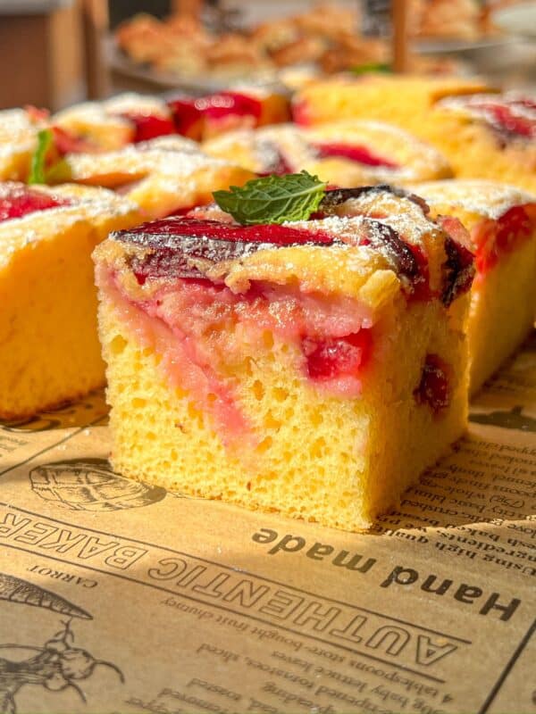 A close-up of a slice of yellow sponge cake topped with red fruit and a dusting of powdered sugar, garnished with a fresh mint leaf, sitting on brown parchment paper with printed text.