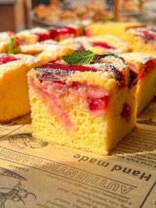 A close-up of a slice of yellow sponge cake topped with fruit pieces, powdered sugar, and a mint leaf, sitting on brown parchment paper with printed text. Other cake slices are visible in the background.