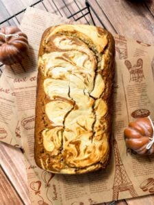A loaf of marbled bread with swirls of light and dark batter sits on parchment paper atop a cooling rack. Decorative mini pumpkins are placed nearby on a wooden surface.