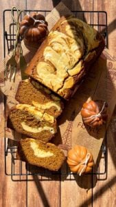 A marble pumpkin bread loaf with cream cheese swirls is sliced on a cooling rack, surrounded by decorative mini pumpkins and gold utensils, with warm sunlight casting soft shadows.