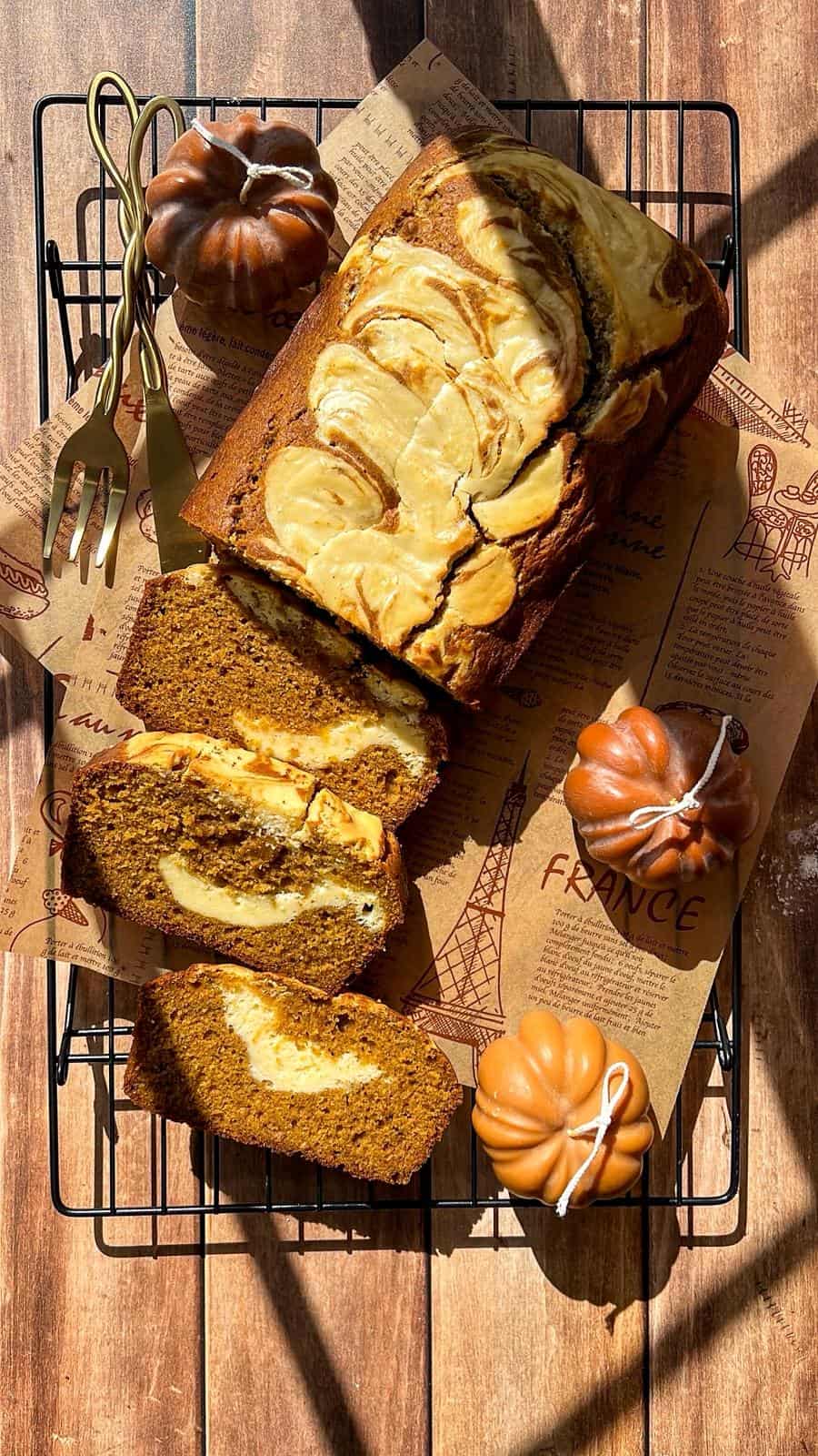 A loaf of pumpkin bread with a cream cheese swirl is sliced on parchment paper atop a cooling rack. Two pumpkin-shaped decorations, a gold fork, and knife are nearby, all on a wooden table in sunlight.