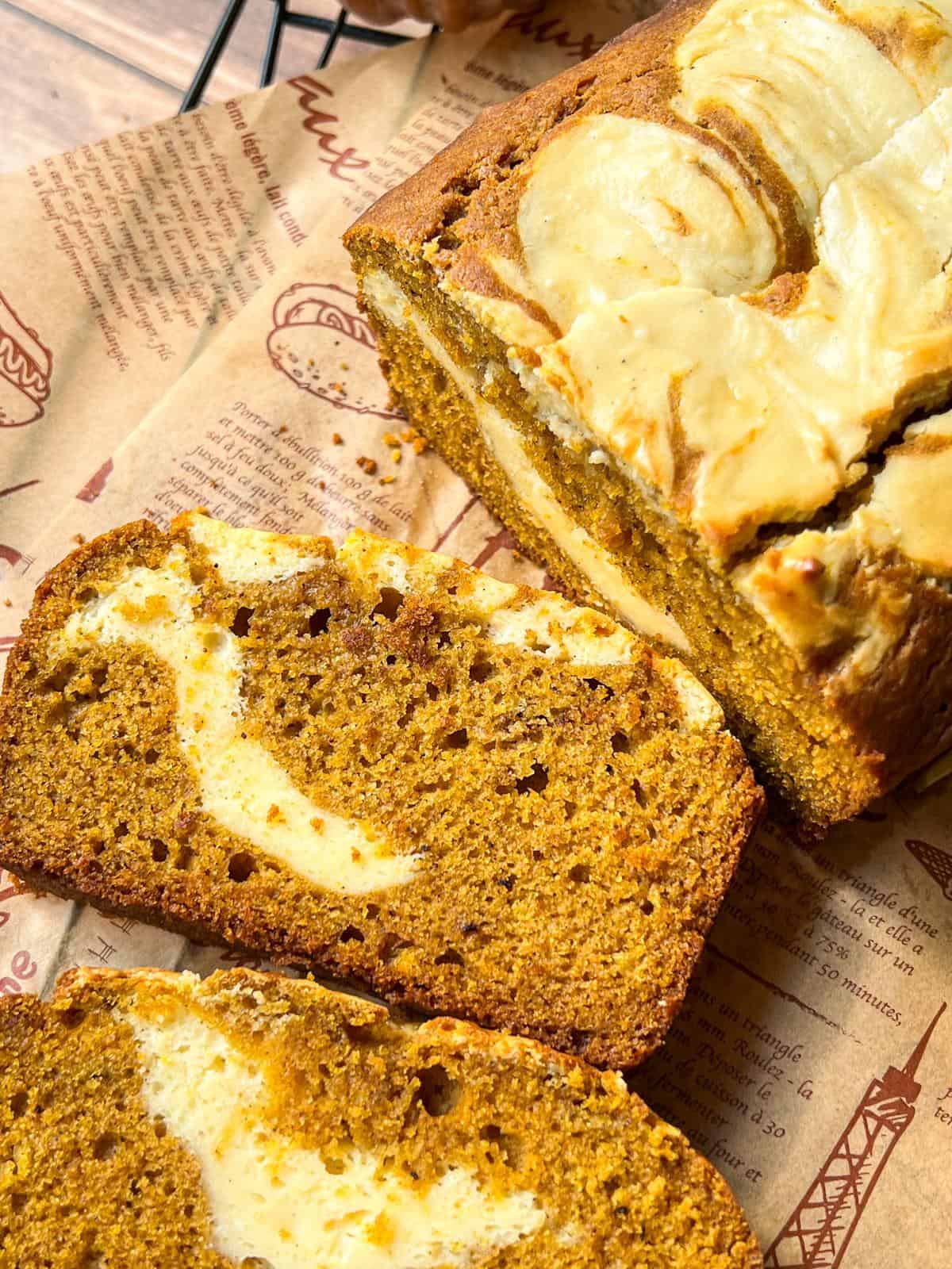 A close-up of sliced pumpkin bread with swirls of cream cheese, placed on brown printed parchment paper. The bread has a moist texture and rich, golden-brown color.