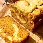 A sliced loaf of pumpkin bread with cream cheese swirls sits on patterned parchment paper, with a small decorative pumpkin in the background. Sunlight highlights the bread’s moist texture and marbled top.