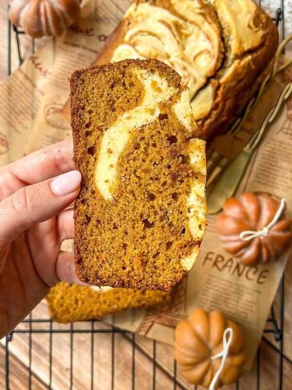 A hand holds a slice of pumpkin bread with a cream cheese swirl; the rest of the loaf sits on a cooling rack, surrounded by decorative mini pumpkins and brown parchment paper.