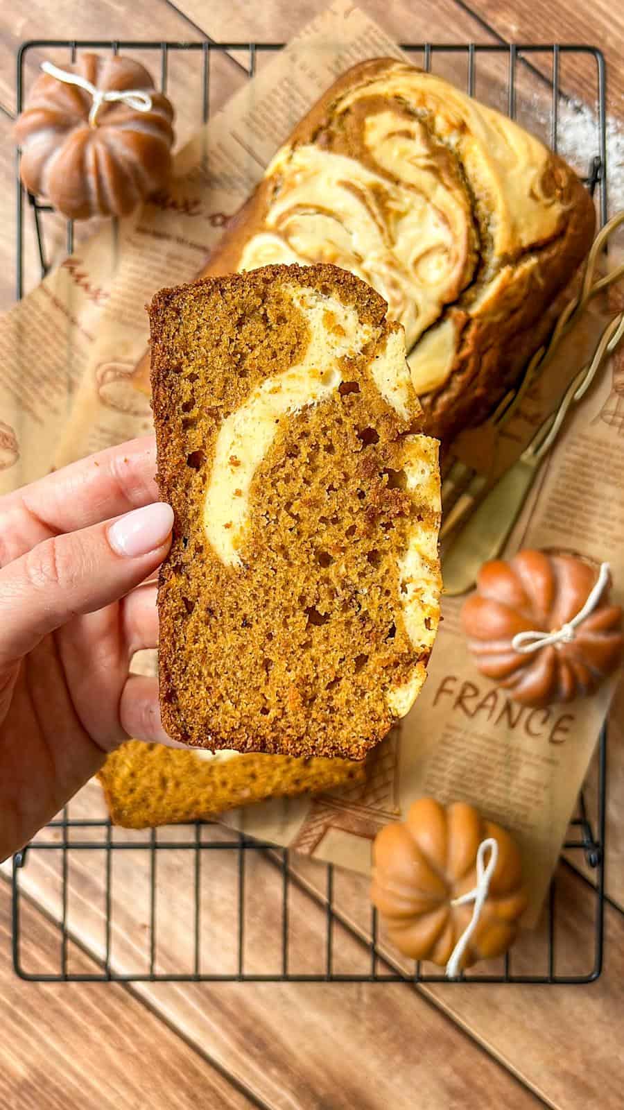 A hand holds a slice of pumpkin bread with a cream cheese swirl; the rest of the loaf sits on a cooling rack, surrounded by decorative mini pumpkins and brown parchment paper.