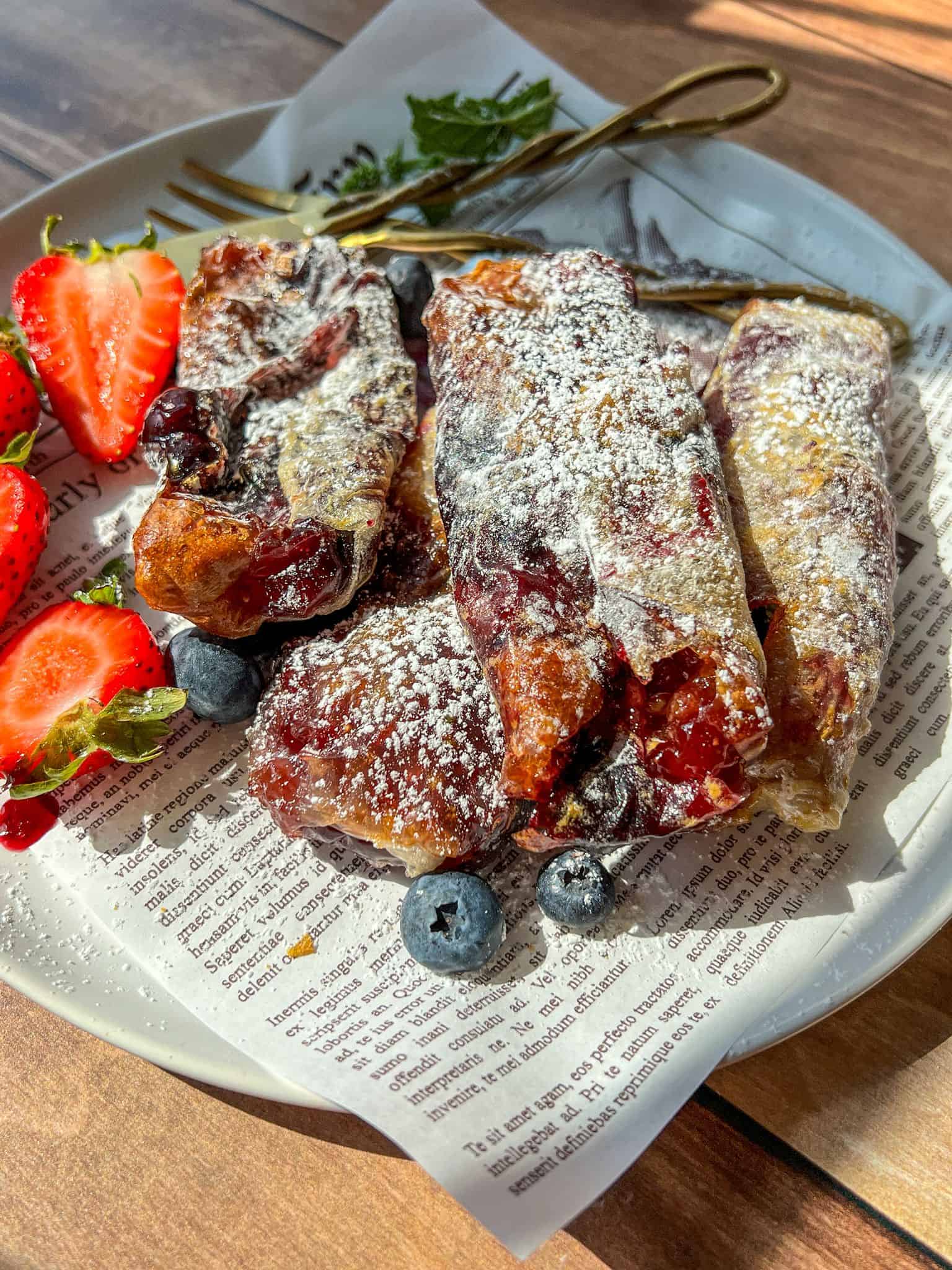 A plate with powdered sugar-dusted pastries, fresh strawberries, and blueberries, served on a piece of printed paper with gold forks beside the food. Sunlight highlights parts of the food and plate.