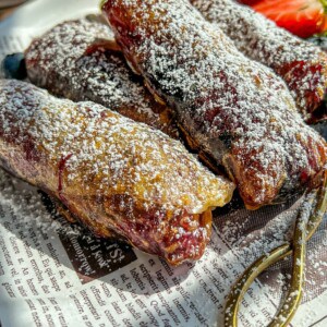 Three powdered sugar-dusted pastries rest on a newspaper-lined plate, with sliced strawberries and a gold fork beside them. Sunlight highlights the flaky texture and golden-brown color of the pastries.