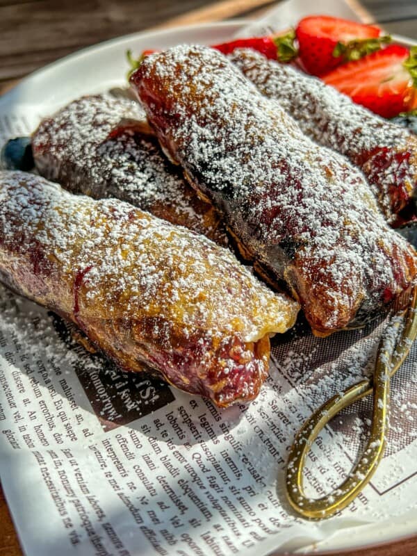 Three powdered sugar-dusted pastries rest on a newspaper-lined plate, with sliced strawberries and a gold fork beside them. Sunlight highlights the flaky texture and golden-brown color of the pastries.