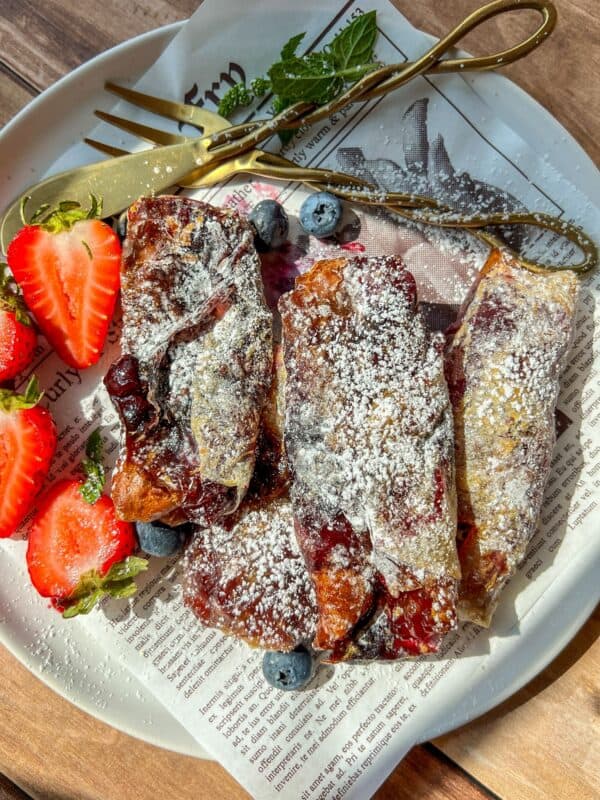 A plate with dessert rolls dusted with powdered sugar, garnished with fresh strawberries, blueberries, and mint, next to a gold fork and knife on a sheet of newspaper-style paper.