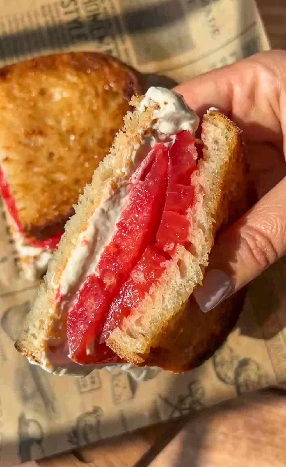 A hand holds a sandwich with thick slices of fresh tomato and a layer of creamy spread between two pieces of toasted bread. The background is a sheet of newspaper-style parchment paper.