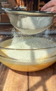 A close-up of flour being sifted through a mesh strainer into a glass bowl filled with a yellow batter, all set on a wooden table with sunlight streaming in.